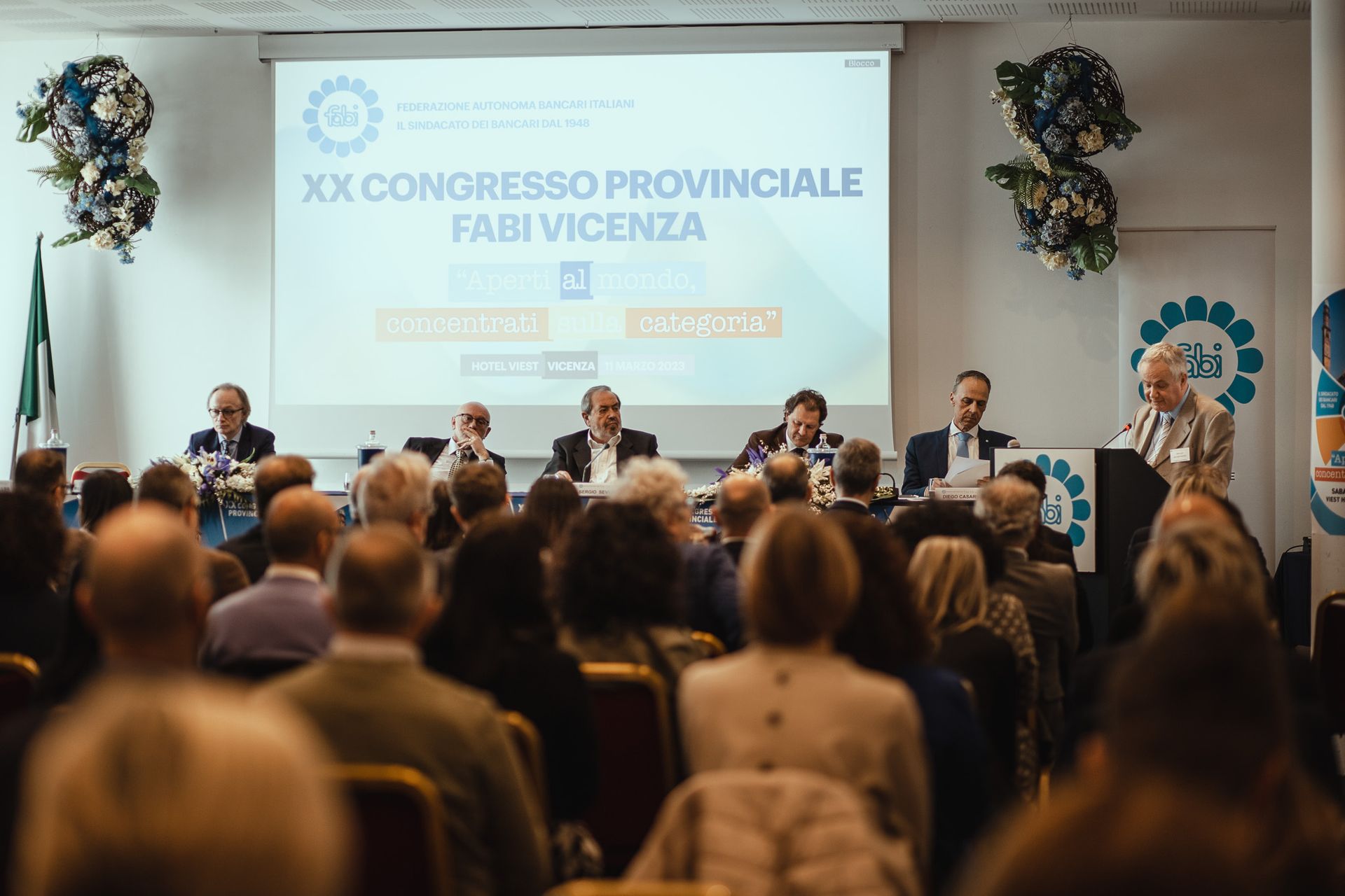 Un folto gruppo di persone è seduto davanti allo schermo di un proiettore durante una conferenza.