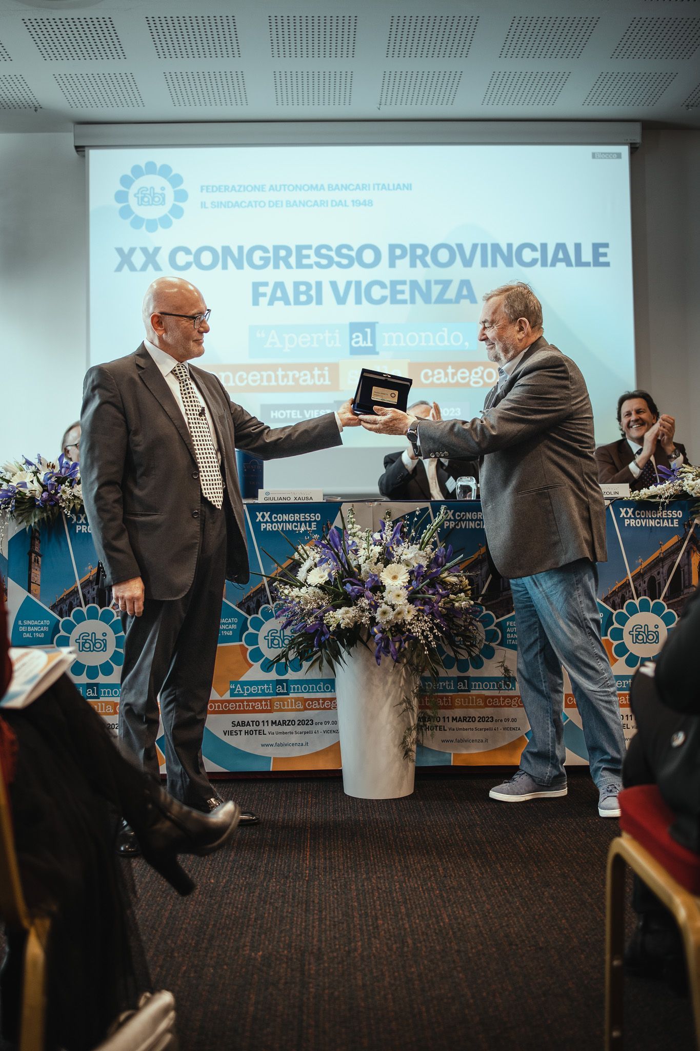 Un uomo in giacca e cravatta consegna una targa a un altro uomo davanti allo schermo di un proiettore.