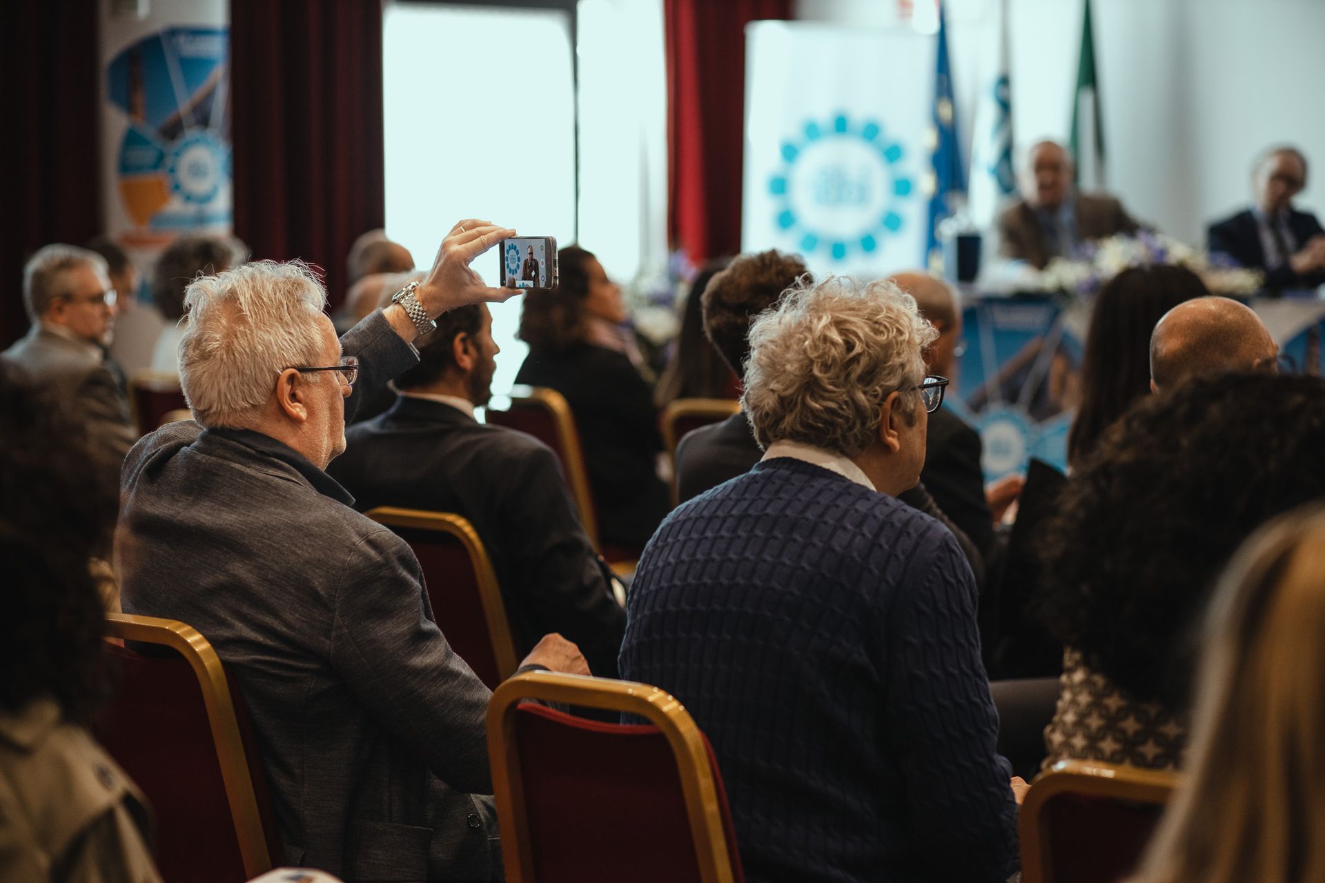 Un uomo sta scattando una foto di un gruppo di persone sedute su alcune sedie durante una conferenza.