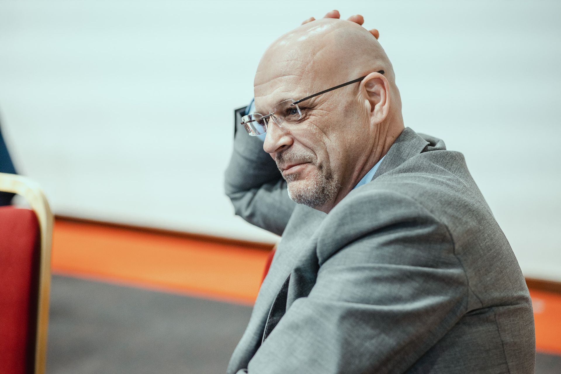 Un uomo calvo con gli occhiali è seduto su una sedia con la mano sulla testa.
