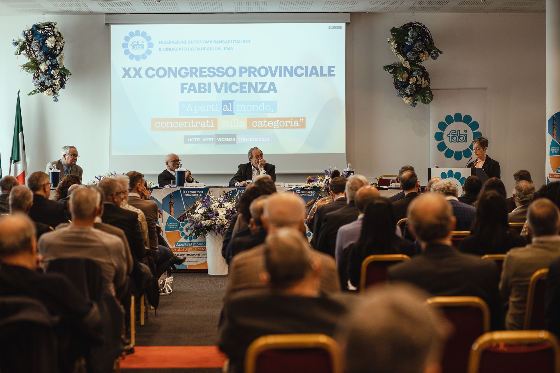 Un gruppo di persone è seduto su delle sedie davanti allo schermo di un proiettore.