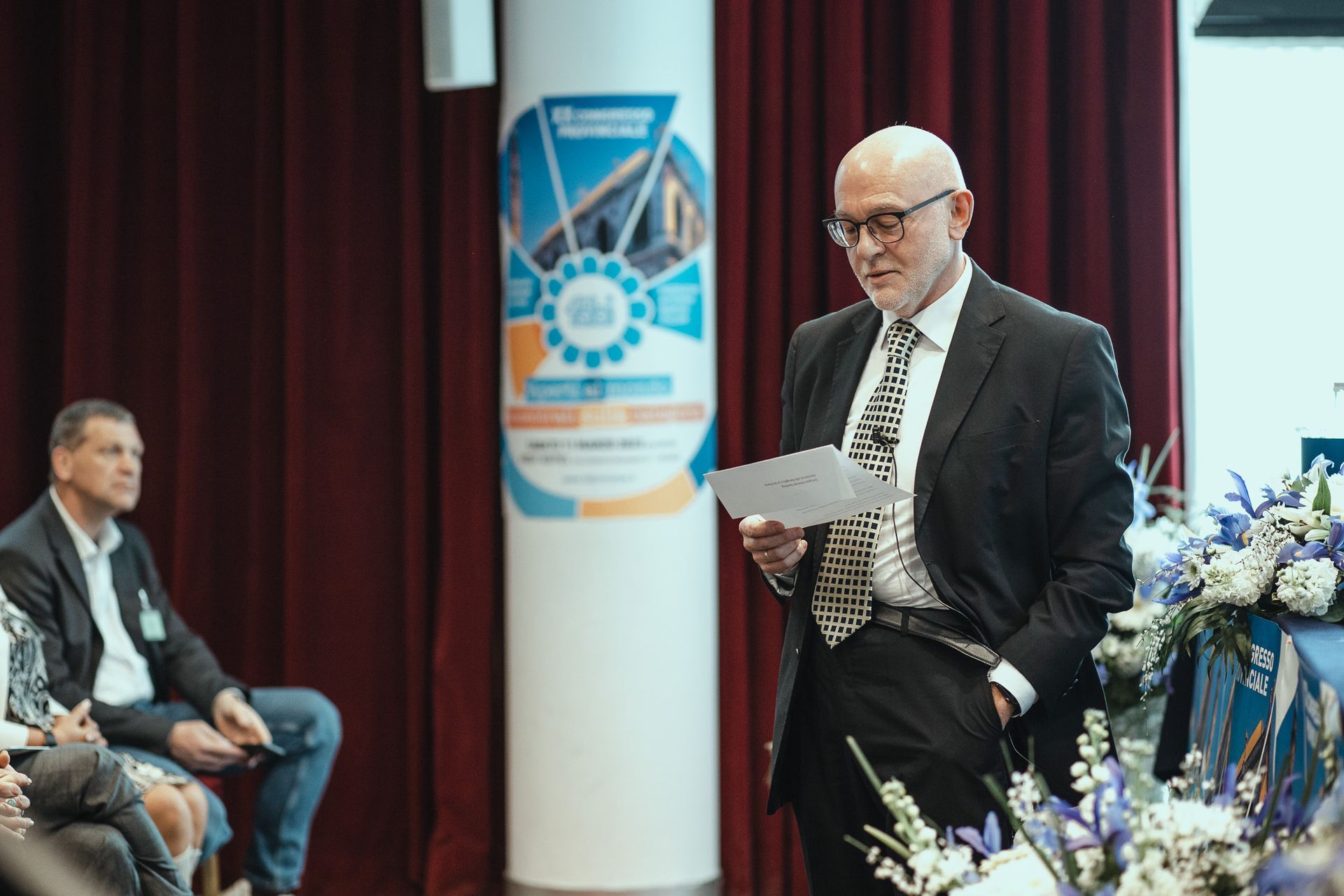 Un uomo in giacca e cravatta tiene un discorso davanti a un gruppo di persone.