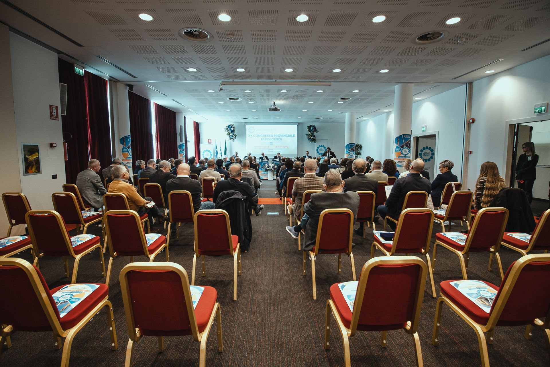 Un folto gruppo di persone è seduto su file di sedie rosse in una sala conferenze.