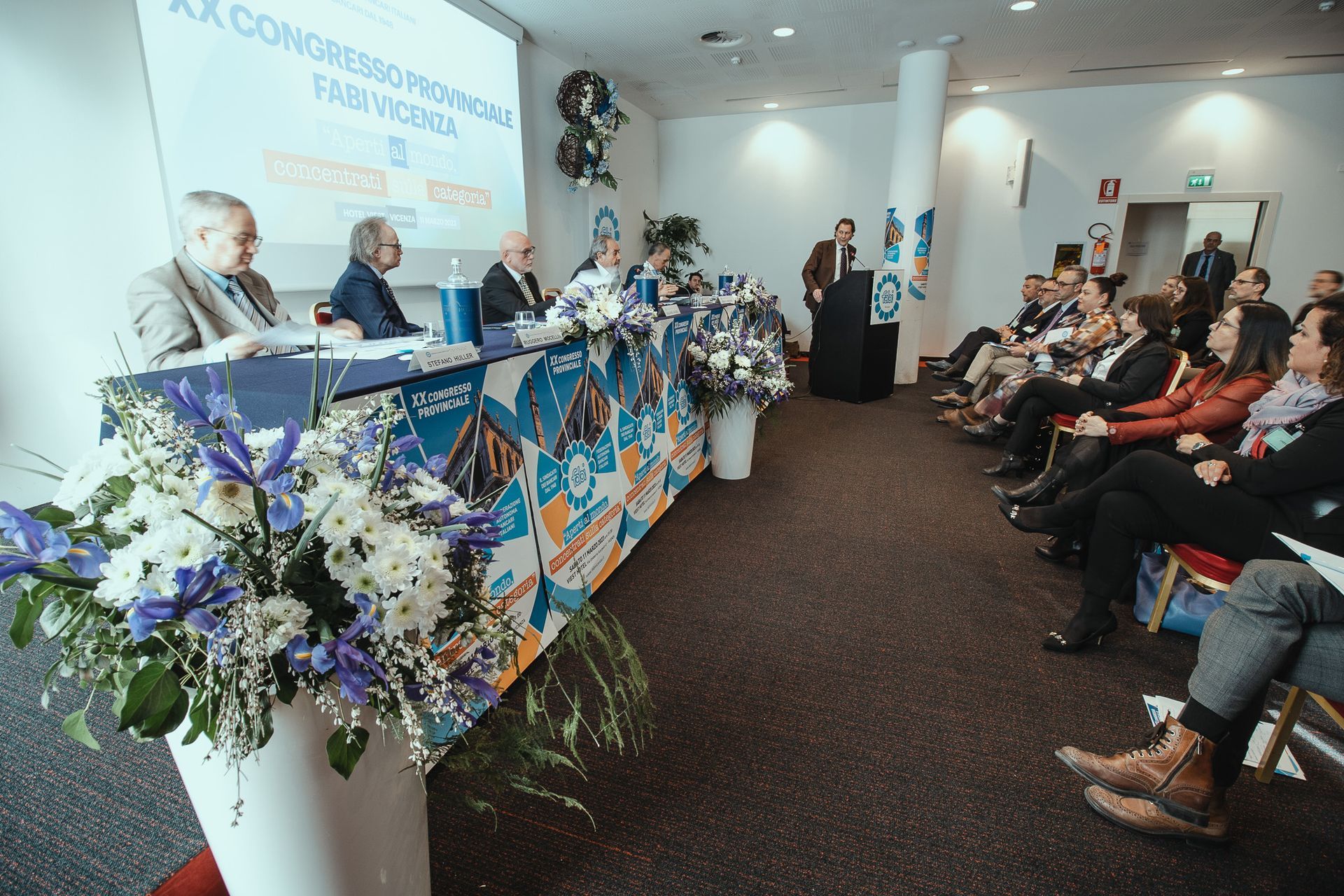 Un gruppo di persone è seduto a un tavolo con dei fiori davanti a sé.