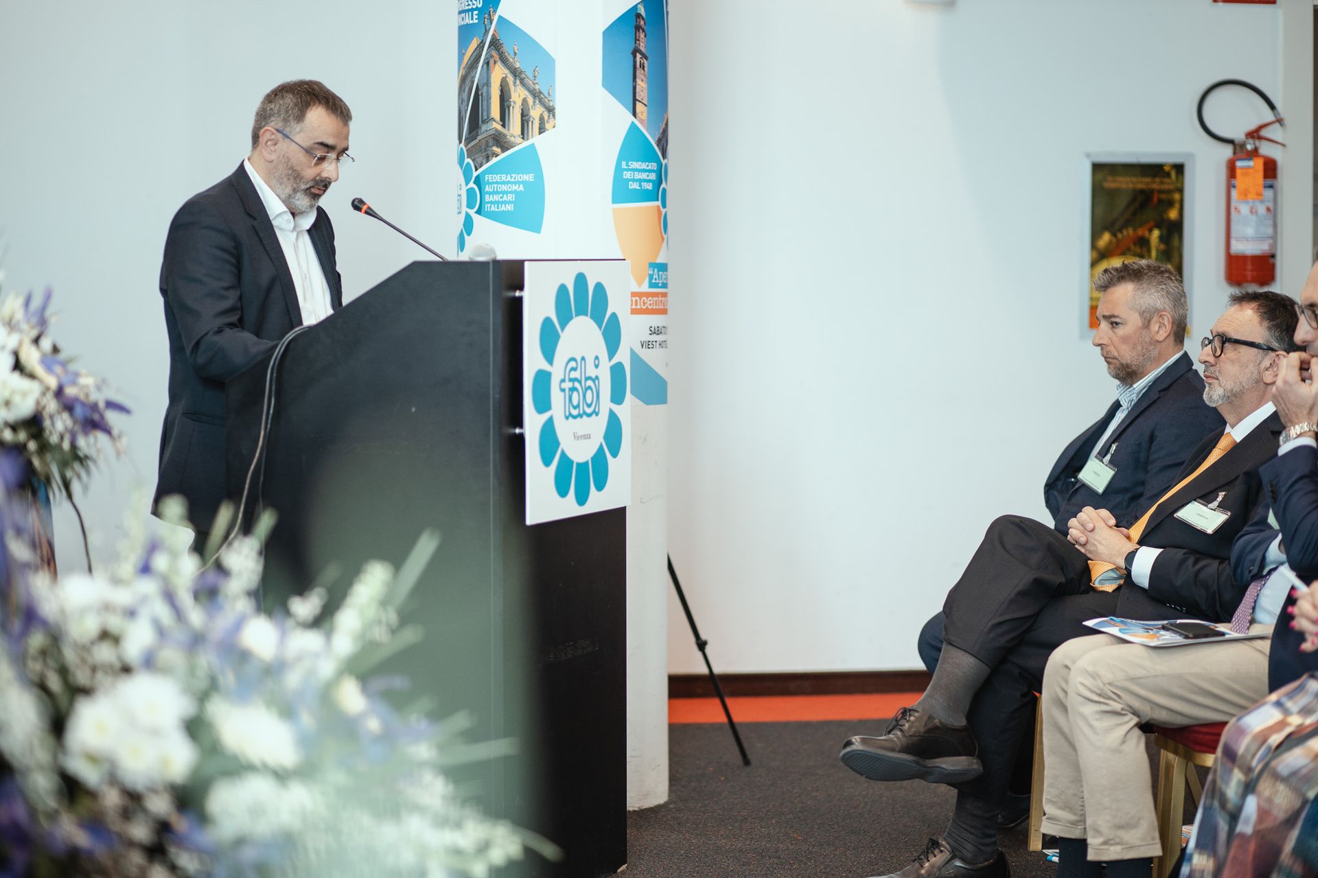 Un uomo tiene un discorso su un podio davanti a un gruppo di persone sedute su sedie.