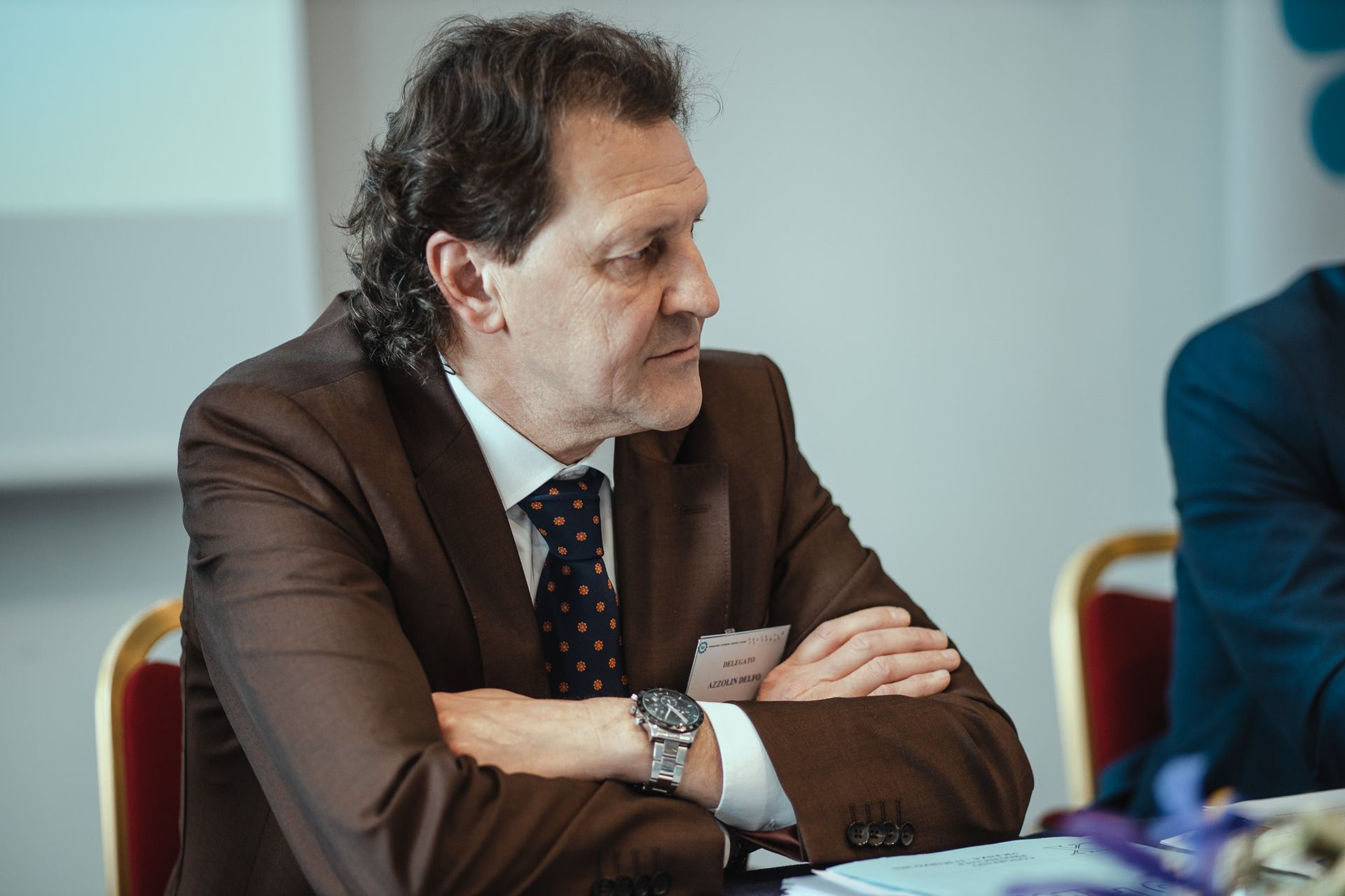 Un uomo in giacca e cravatta è seduto a un tavolo con le braccia incrociate.