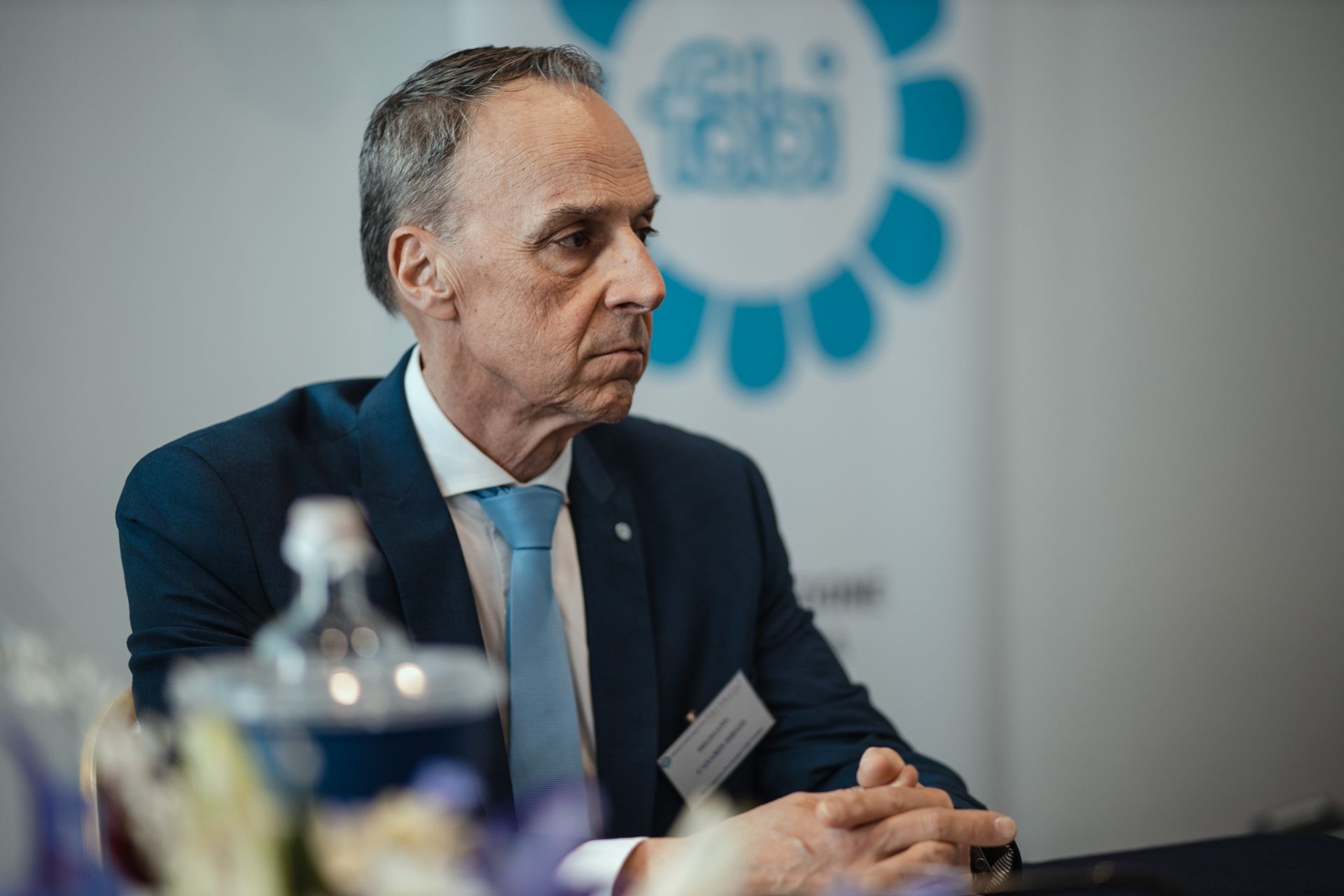 Un uomo in giacca e cravatta è seduto a un tavolo con le mani giunte.