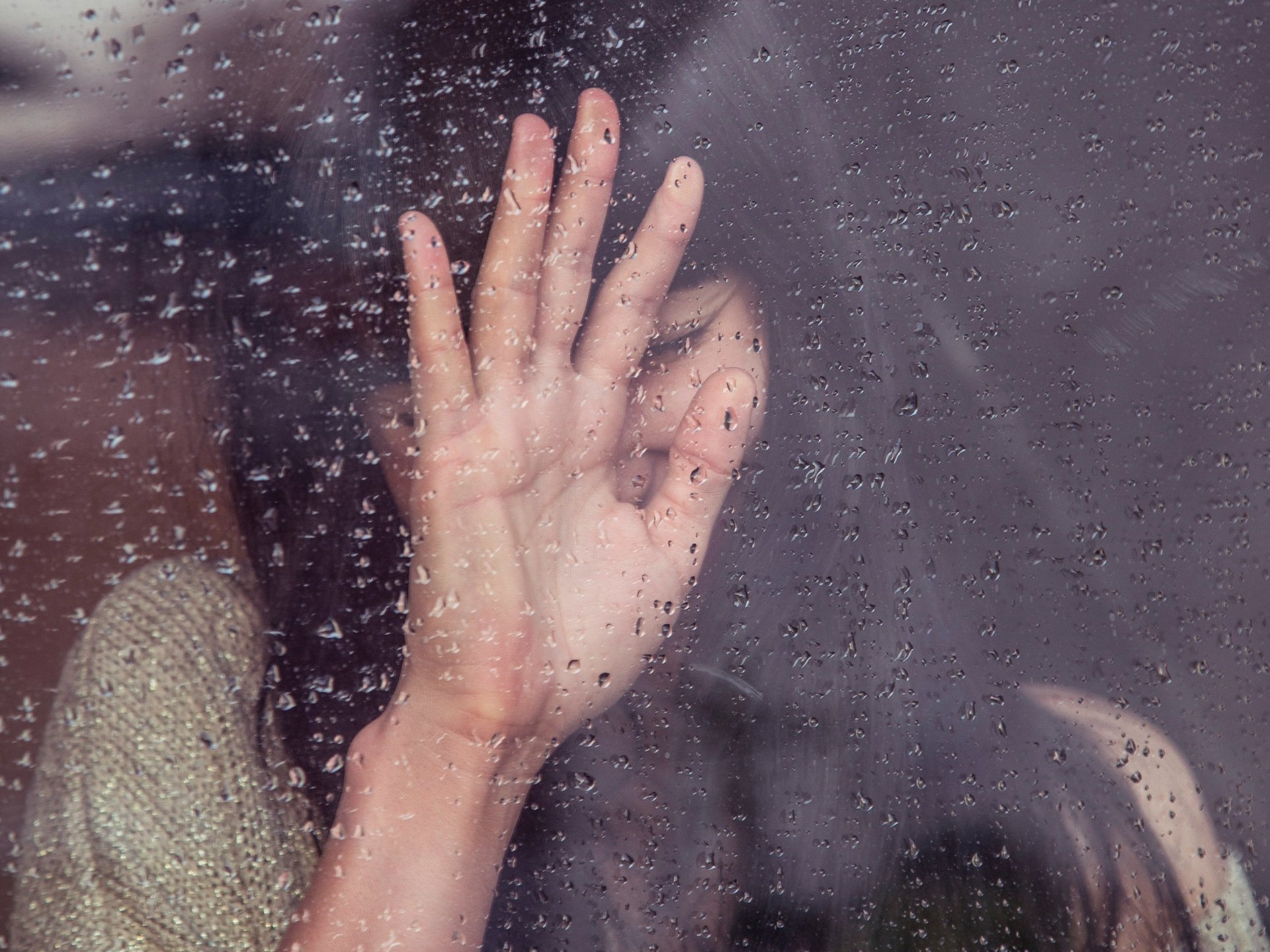 Una donna si copre il viso con la mano dietro una finestra su cui cadono gocce di pioggia.