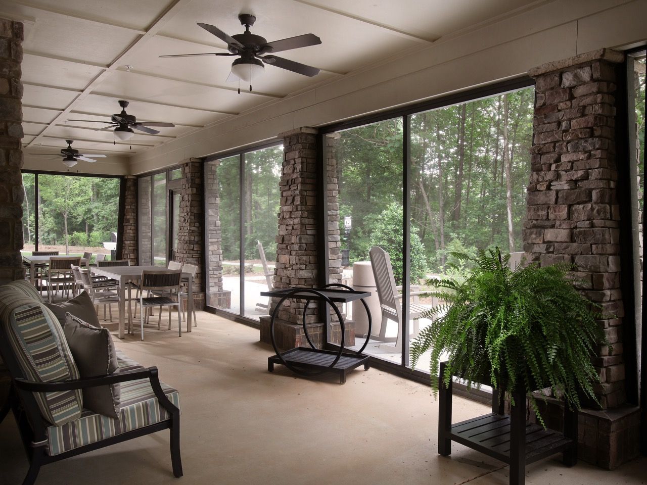 Covered outdoor lounge with stone columns, ceiling fans, and glass walls overlooking trees.