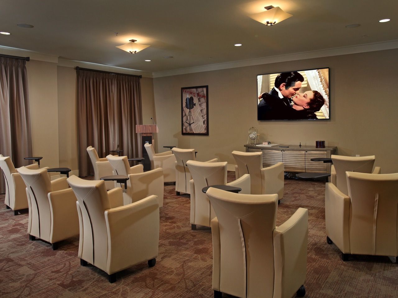 Communal lounge with beige upholstered chairs arranged in rows facing a wall-mounted TV.