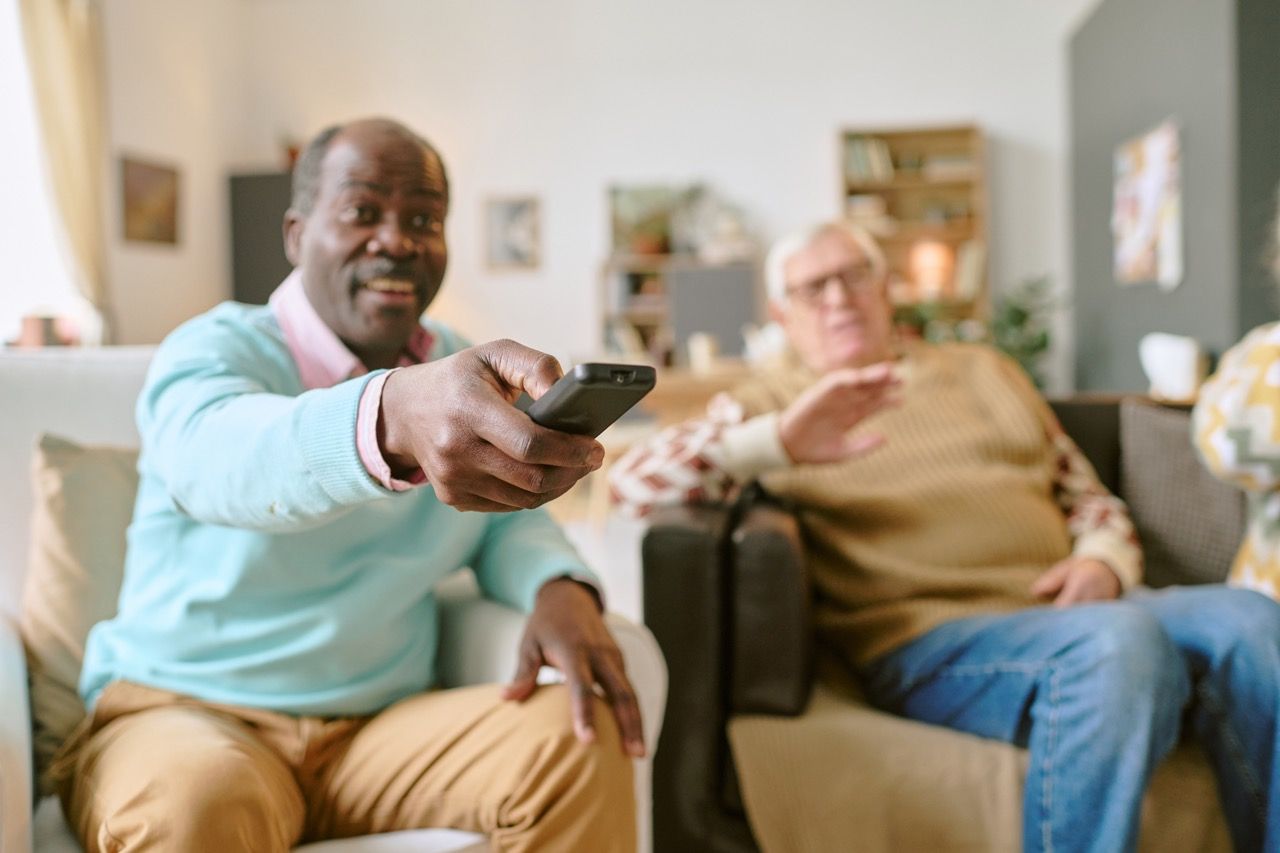 Two men on a sofa; one holding a TV remote, reaching forward. The other gestures with his hand. Living room.