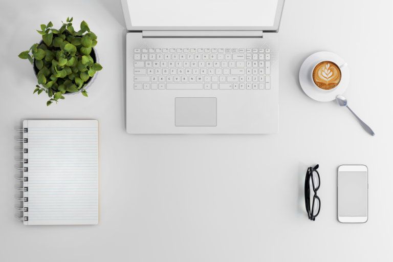 A white desk with a laptop, cell phone, glasses , and a cup of coffee.