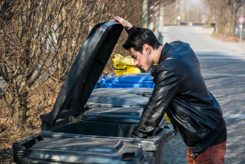 A man in a leather jacket is looking into a trash can.