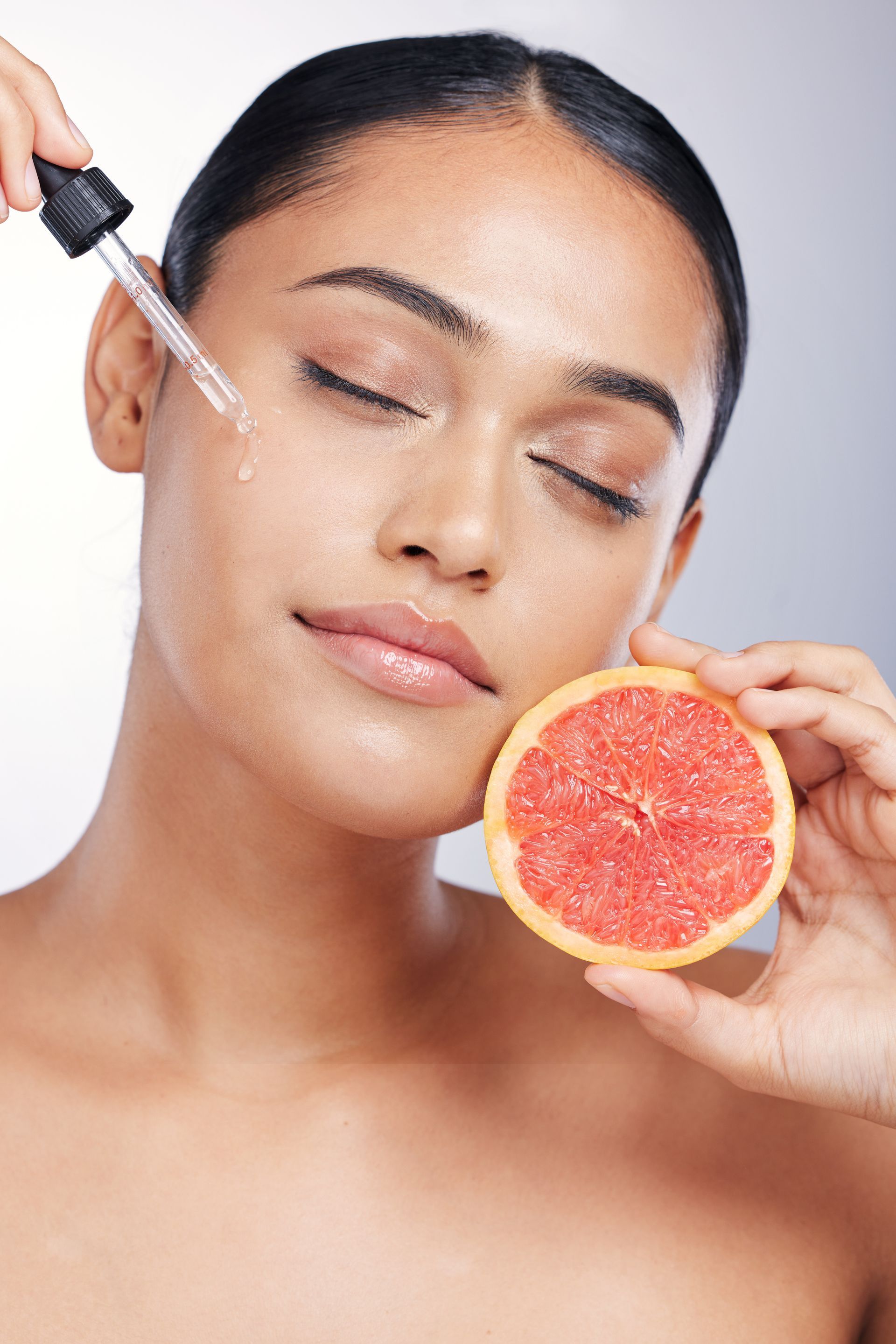 Eine Frau trägt ein Serum auf ihr Gesicht auf, während sie eine Grapefruitscheibe in der Hand hält.