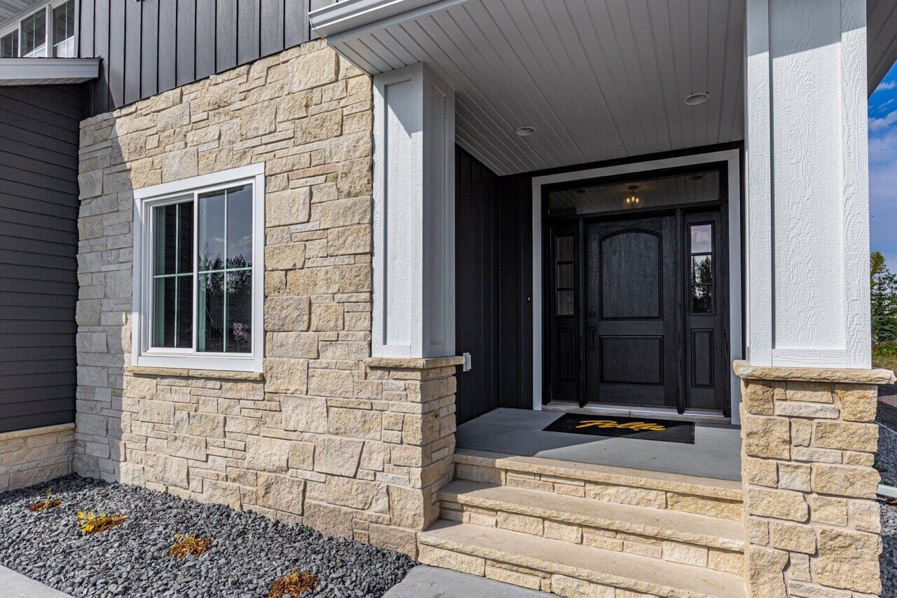 A modern house entrance featuring a beige stone exterior, dark door, white porch columns, and gray gravel landscaping.
