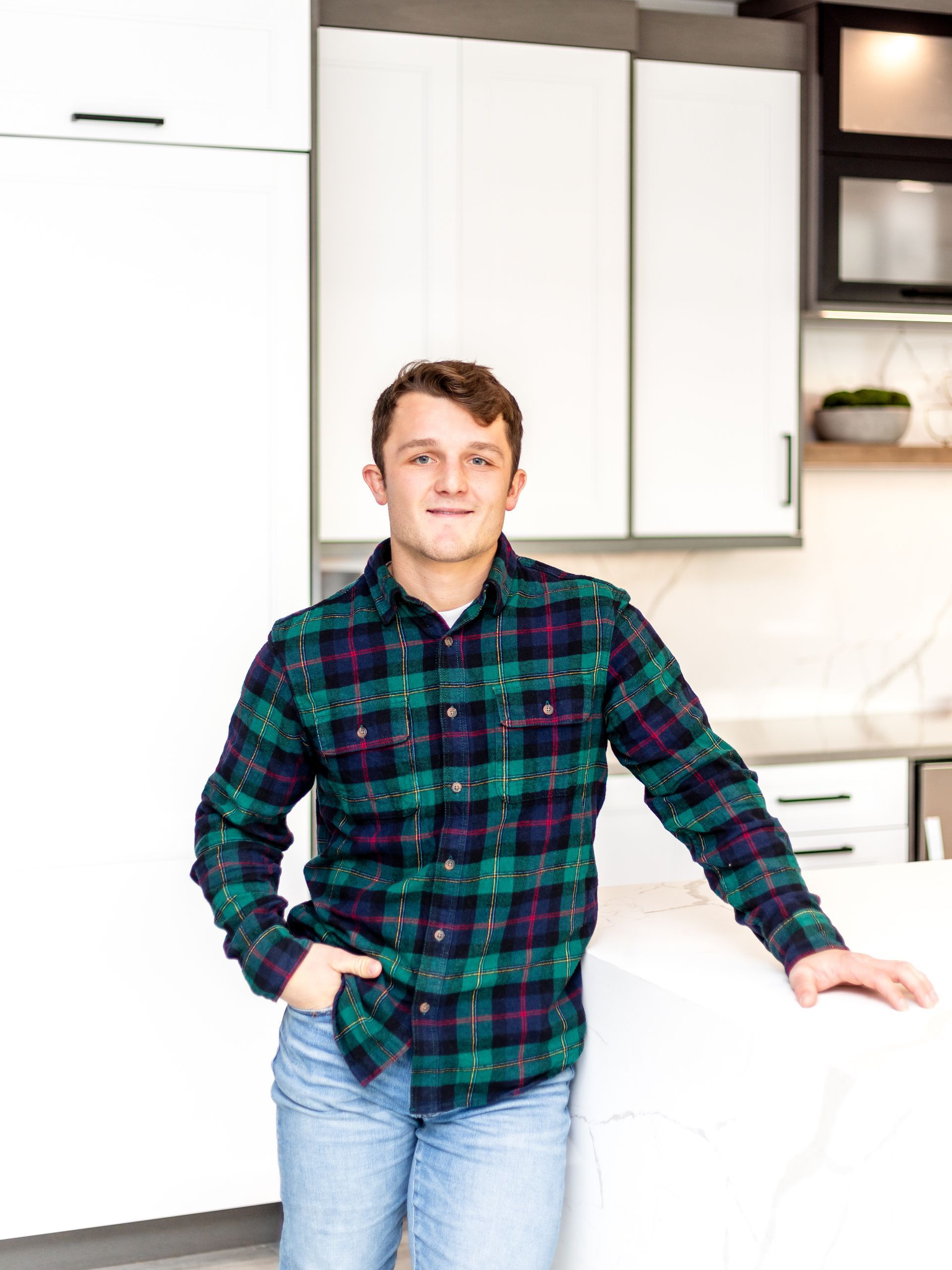 Man in plaid shirt and jeans, leaning against a white countertop in a modern kitchen.