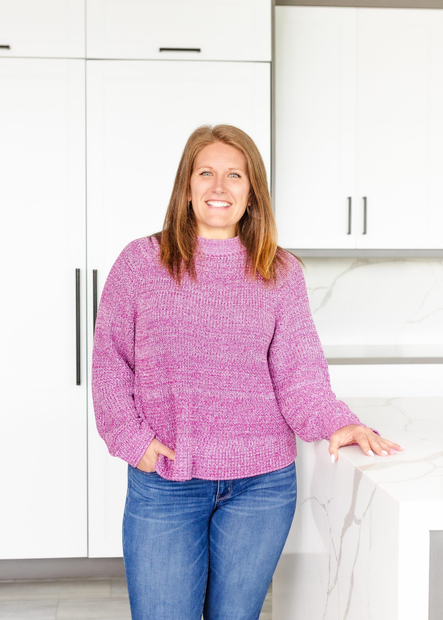 Woman wearing a pink sweater and jeans smiles, leaning on a white countertop in a bright kitchen.