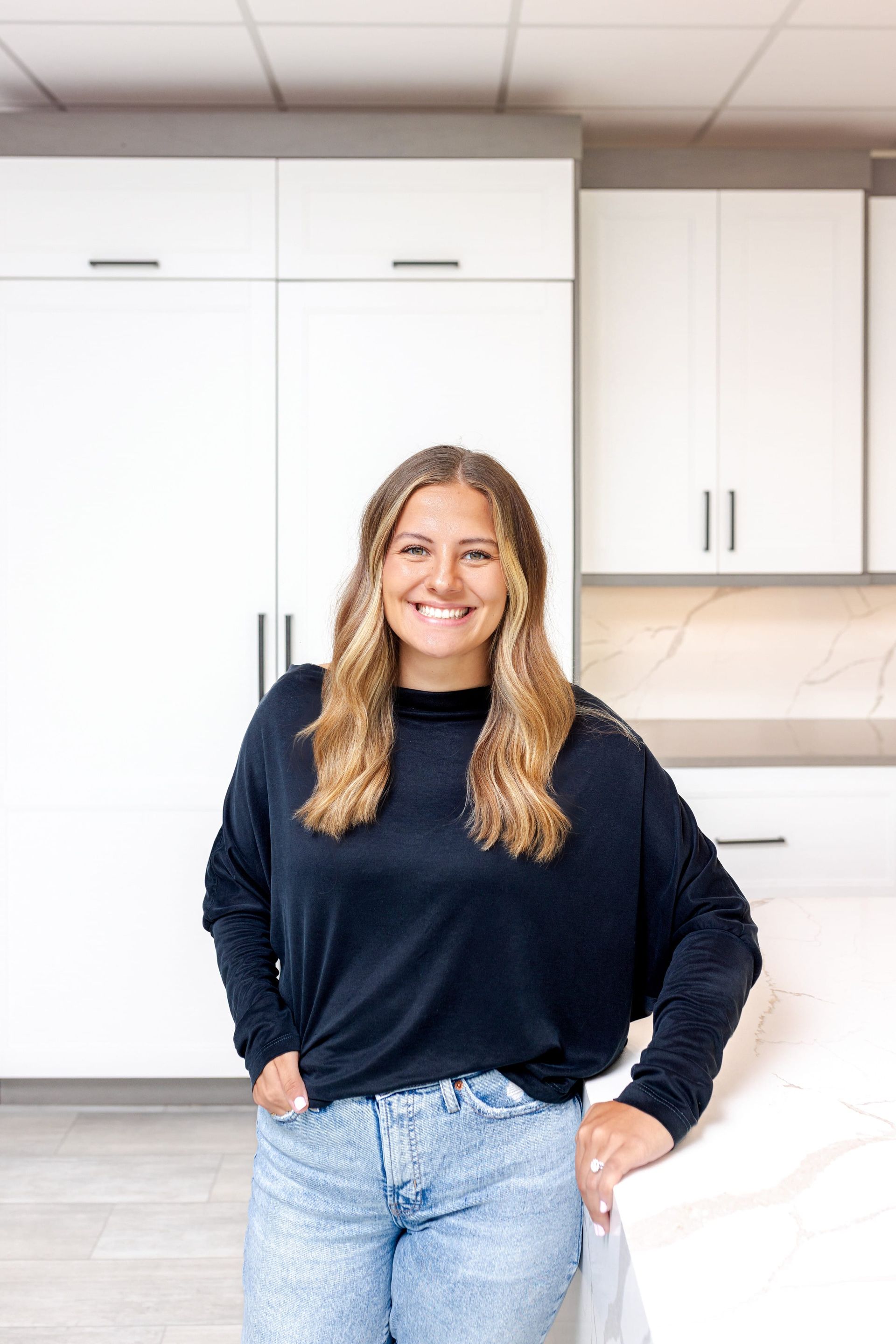 Woman in a kitchen smiling, wearing a black shirt and jeans, leaning on a white countertop.