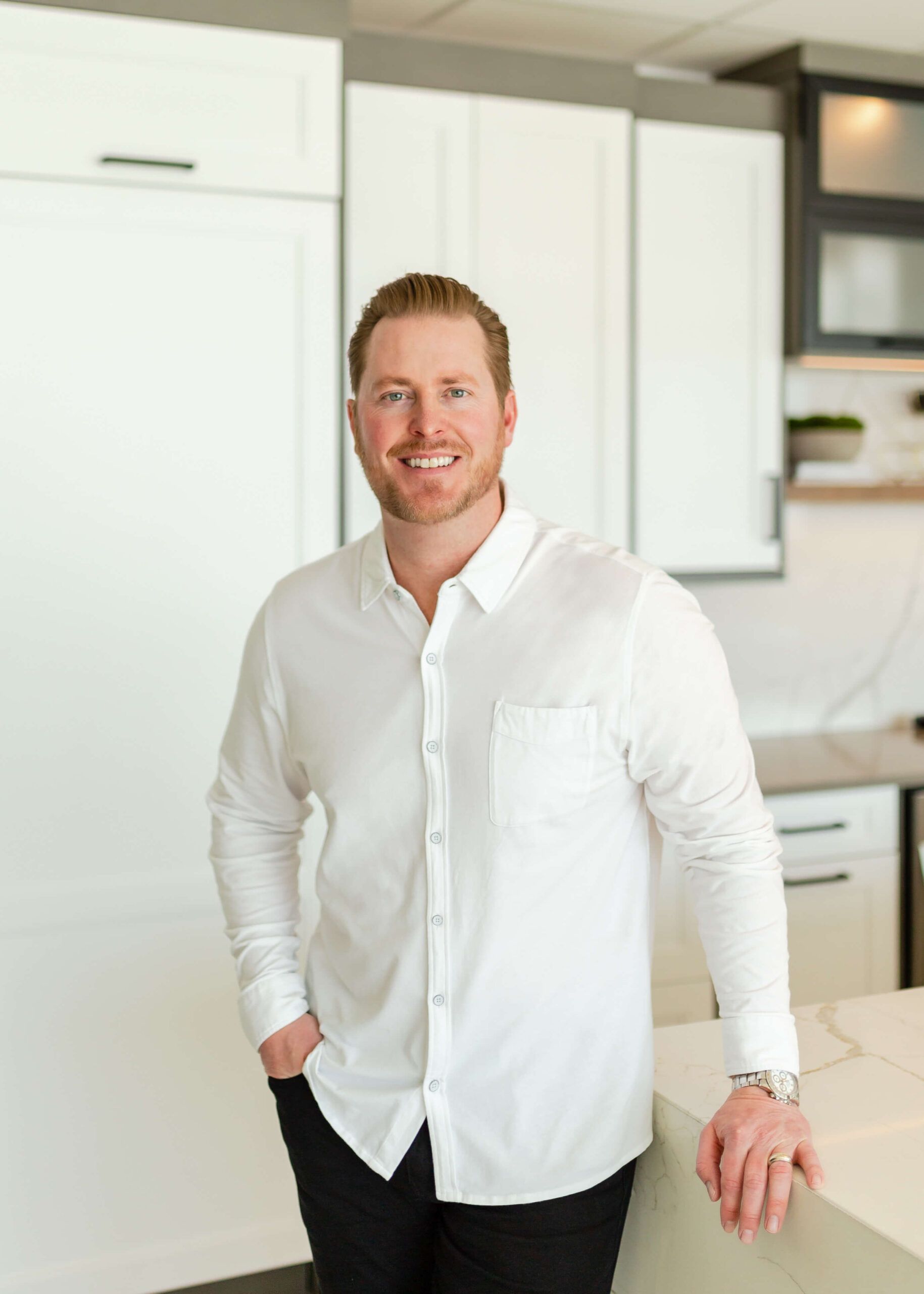Man in white shirt smiling in a modern kitchen. Leaning on counter with hand in pocket.