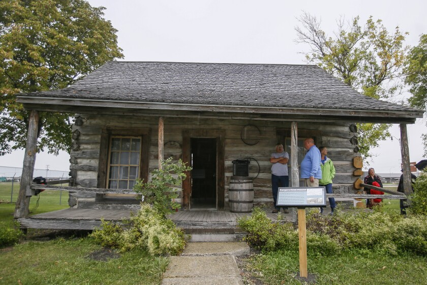 A rustic log cabin with a wooden porch and shake roof, featuring a small barrel and a sign standing in the grassy yard.