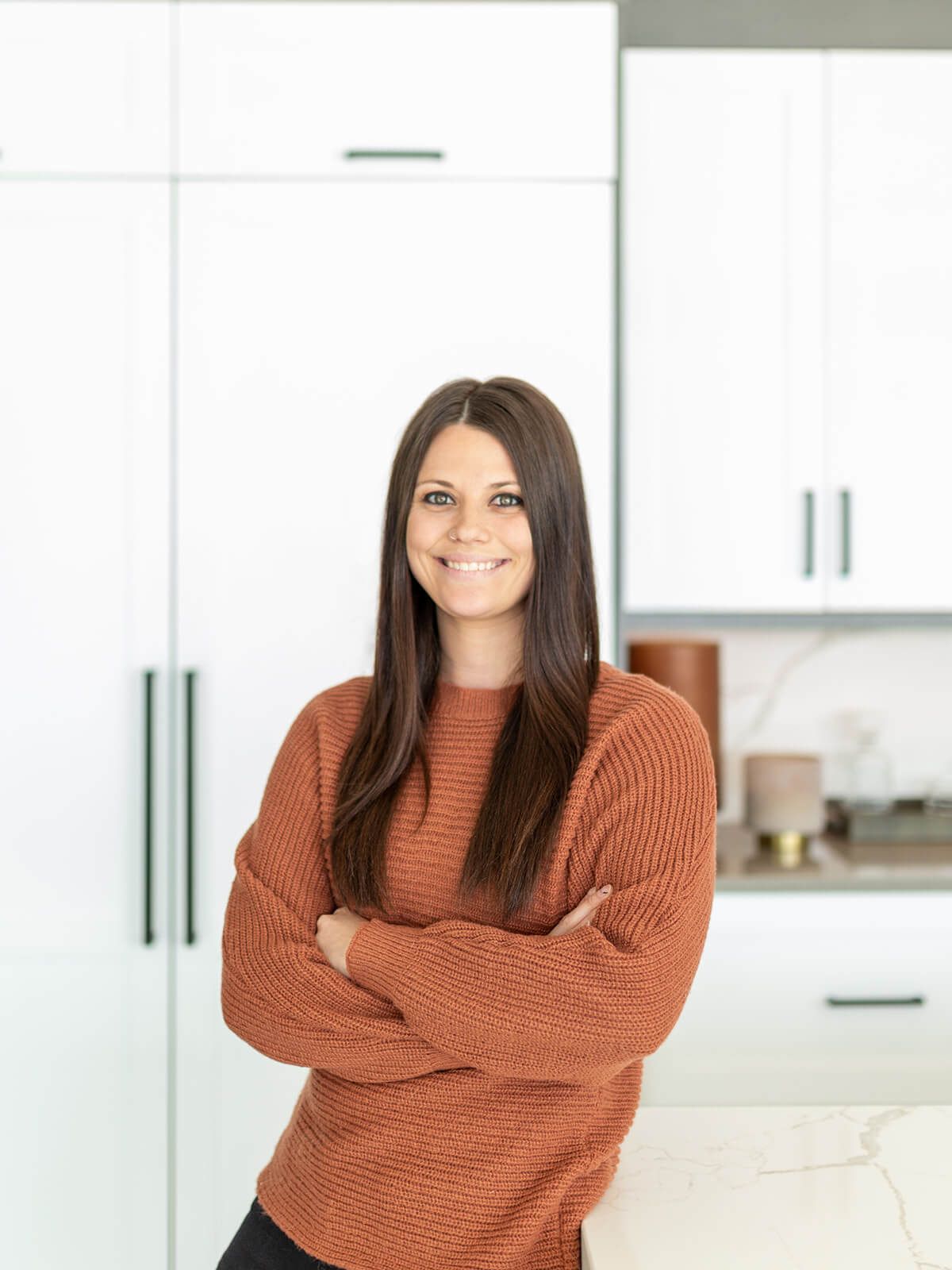 Woman in orange sweater smiles, arms crossed, in a modern white kitchen.
