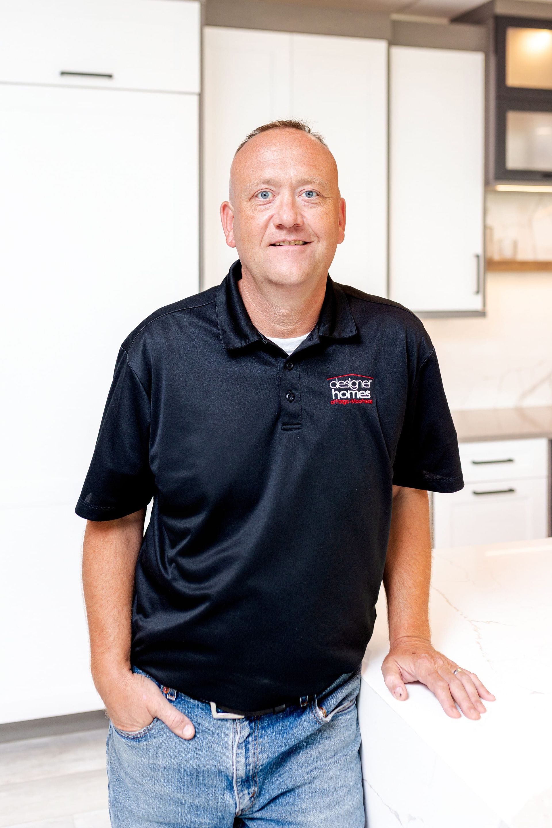 Man in black polo shirt stands in kitchen, leaning on countertop.