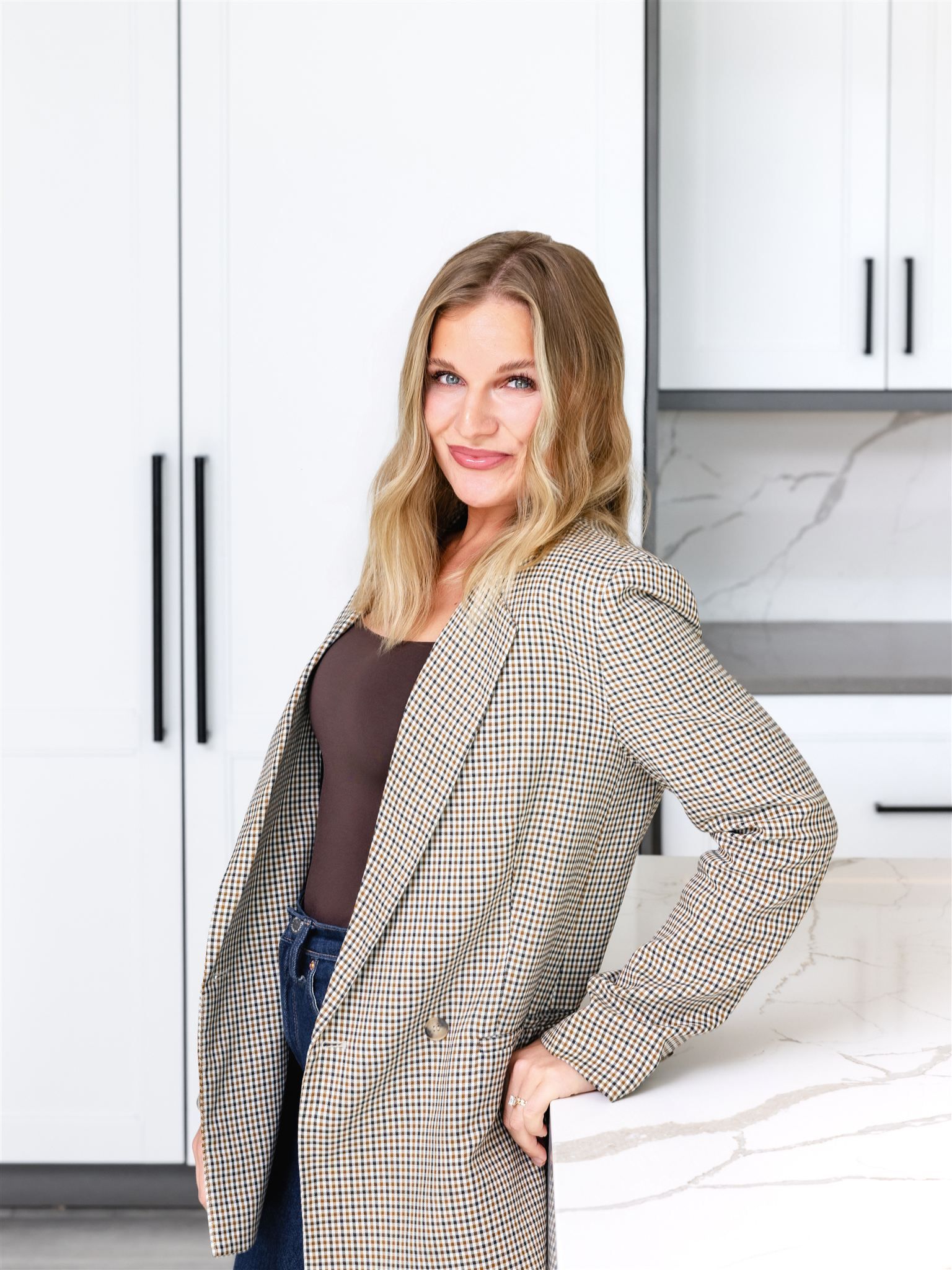 Woman in plaid blazer, leans on a kitchen counter with neutral-toned cabinets and marble surface.
