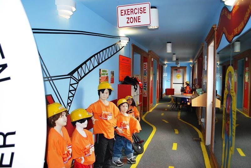 Four children in orange shirts and yellow hard hats stand in a hallway painted with a construction crane theme.
