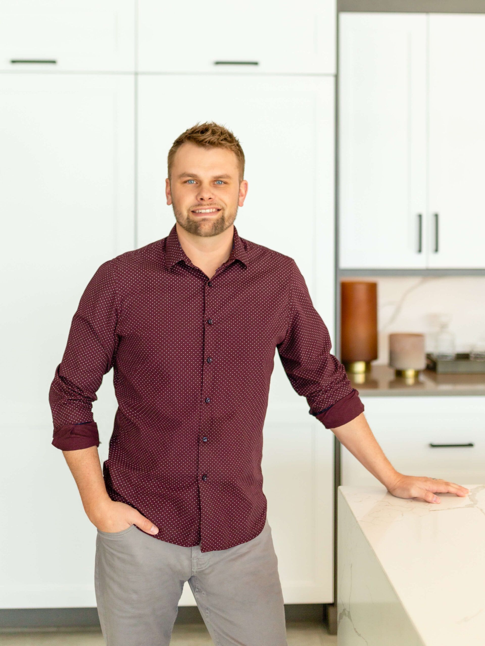 Man in burgundy shirt and gray pants leans on a kitchen countertop.
