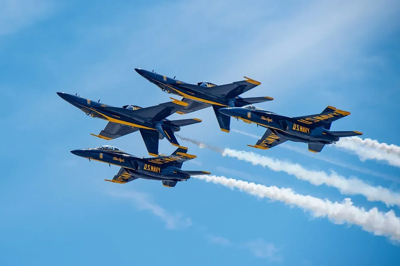 Four Blue Angels F/A-18 Hornet jets flying in a tight formation against a bright blue sky, leaving long white smoke trails.