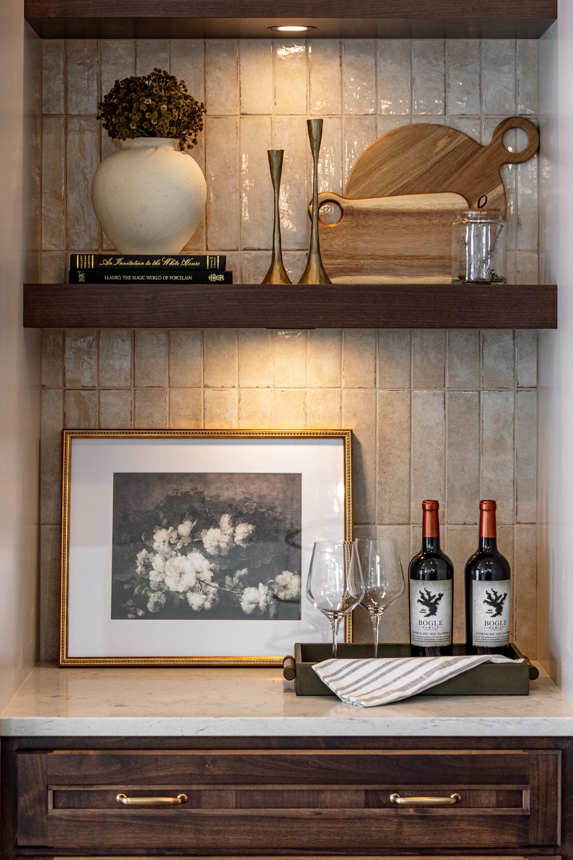 Dark wood shelving with decorative items: vase, candlesticks, cutting board, framed floral art, wine bottles, and glasses.