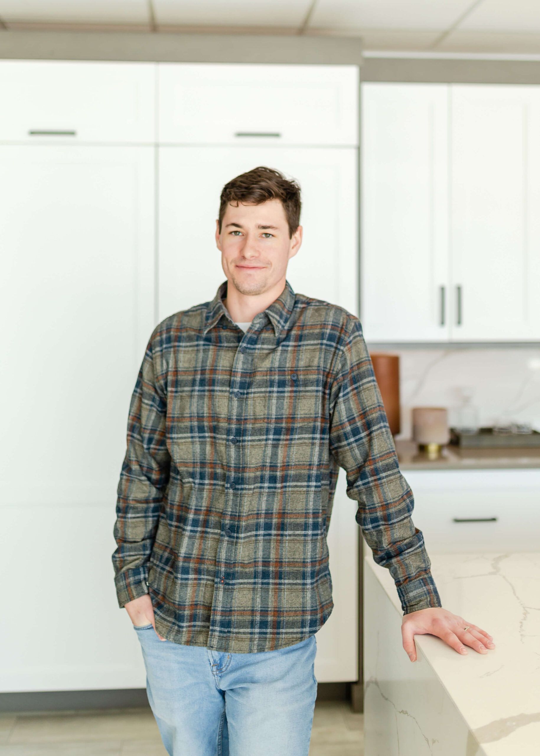 Man in plaid shirt and jeans leans on a kitchen countertop.
