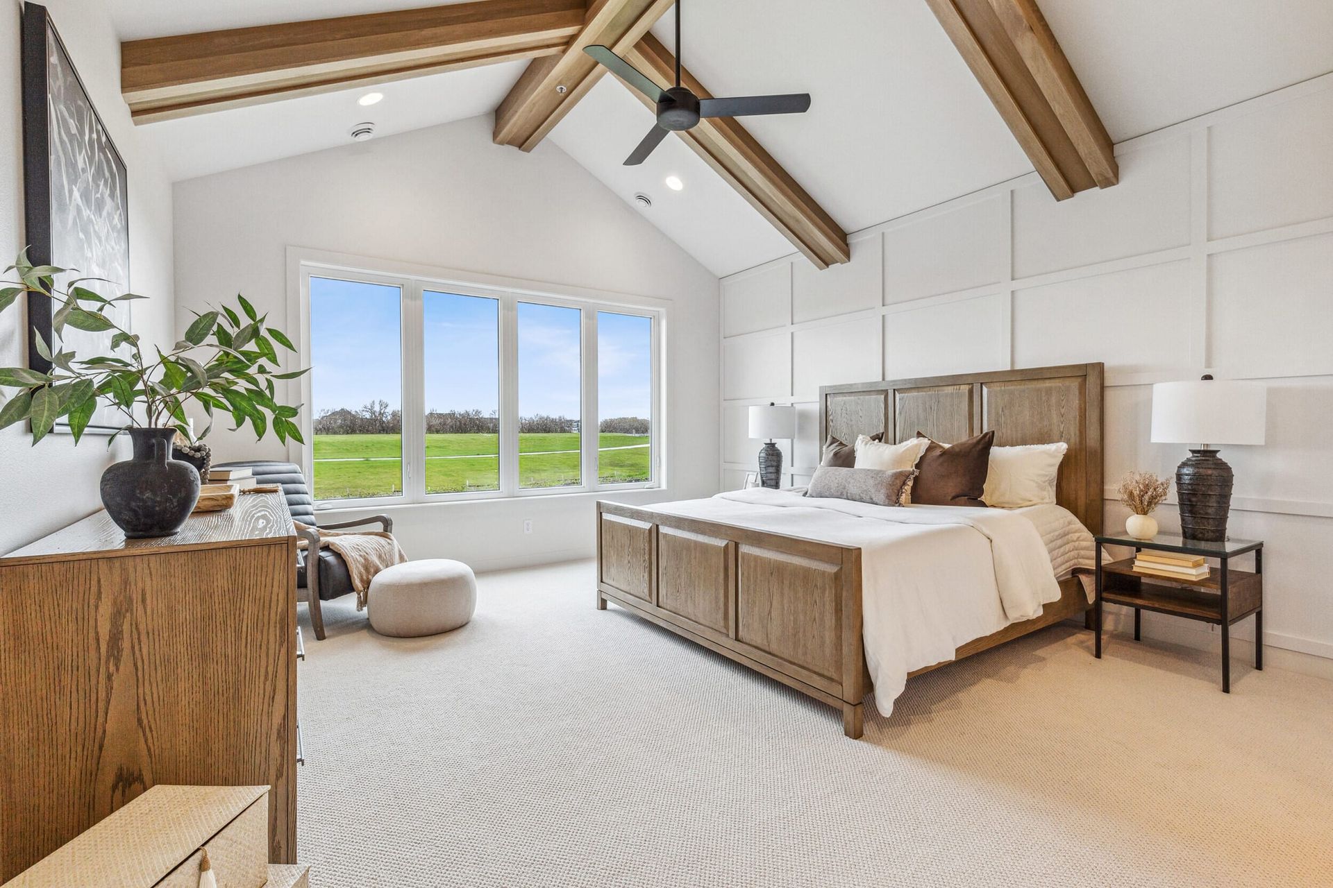 Spacious bedroom with vaulted ceiling, wooden beams, large window overlooking a green field, and a wooden bed.