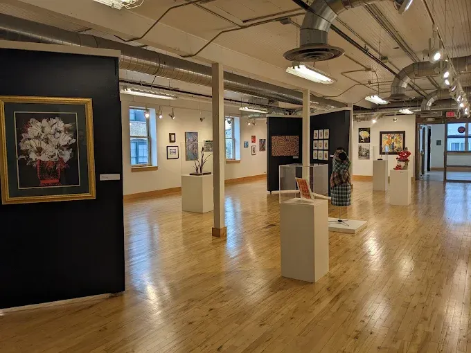 An art gallery with hardwood floors, white pedestals, and various framed artworks displayed on walls and stands.