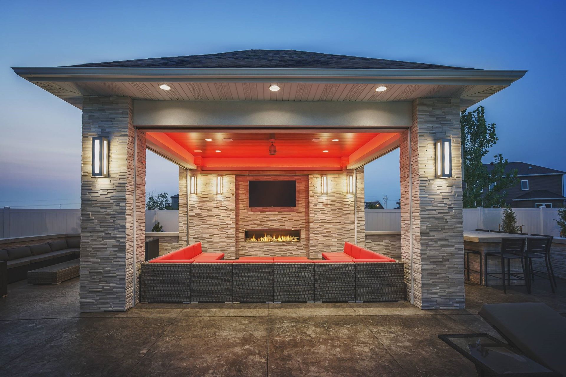 Outdoor patio gazebo with fireplace, TV, seating, and lighting at dusk.