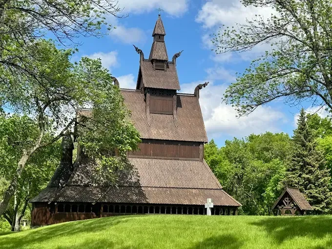 The Hopperstad Stave Church replica features dark wood, layered roofs, and dragon head gables on a grassy hill under sky.