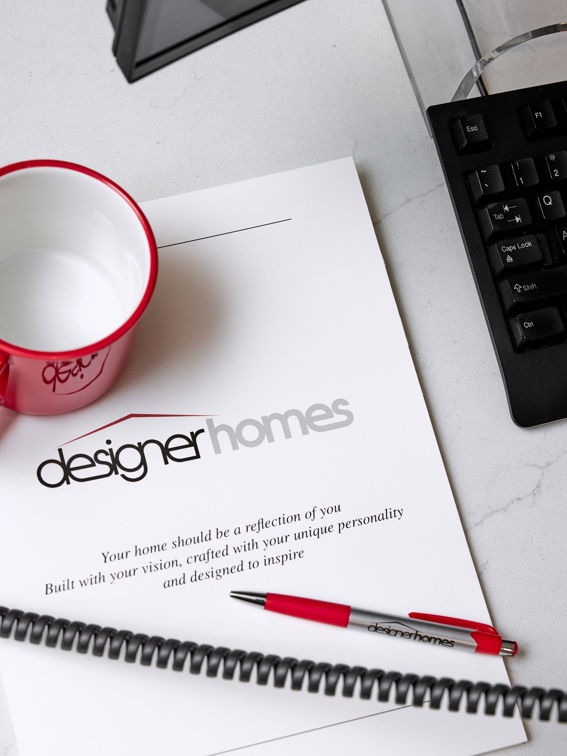 Desk with a notebook, red mug, keyboard, and red pen over a “designer homes” magazine page.