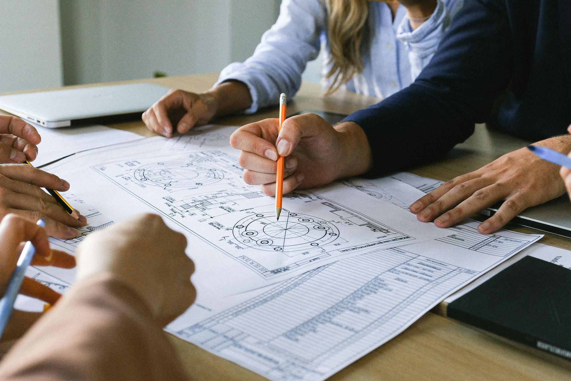 People collaborating around blueprints, using pencils and tablet on a wooden table.