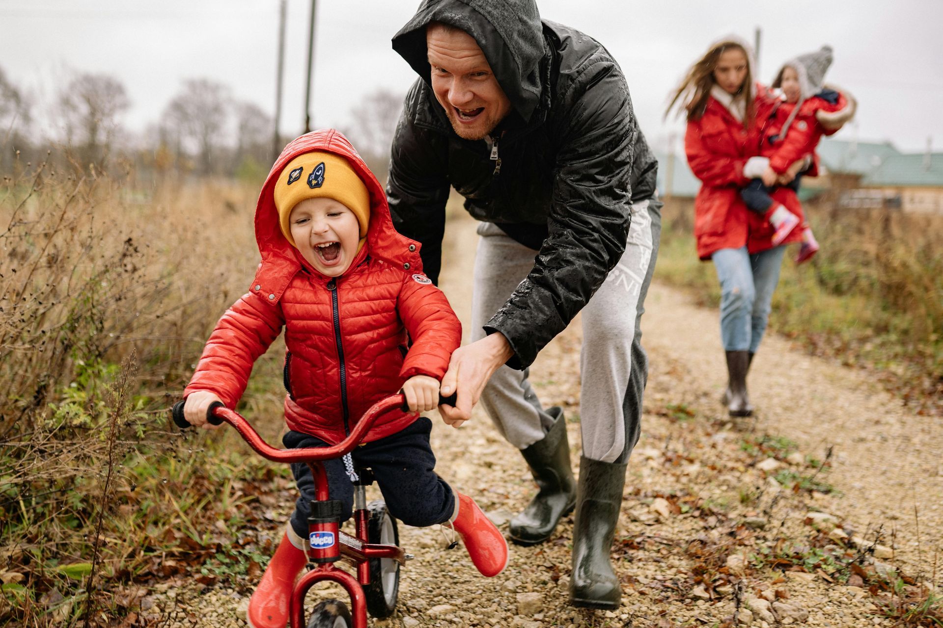A smiling child on a red balance bike is pushed by an adult along a gravel path, with another person holding a toddler nearby.
