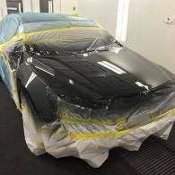 A dark-colored car in a paint booth, covered in protective plastic sheeting and yellow masking tape for refinishing.