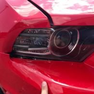 A person's finger points to a gap between the bumper and headlight assembly on a red car, showing damage or misalignment.