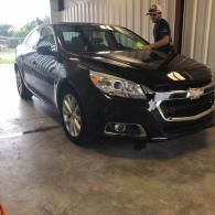 A person wearing a cap polishes the windshield of a shiny black sedan inside a garage.