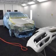 A white car, partially masked with blue plastic and yellow tape, sits in a paint booth next to its unattached bumper.