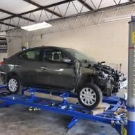 A gray sedan missing its front bumper is mounted on a blue hydraulic lift inside an auto repair shop.