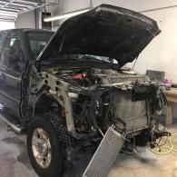 A dark pickup truck with its hood open, missing the front bumper and grille, with a radiator hanging from the front.
