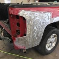 Red pickup truck bed in a garage, featuring a sanded body repair area around the missing taillight.
