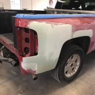 A red pickup truck bed in an auto body shop with body filler applied to the rear panel for repairs.