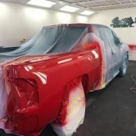 A bright red pickup truck partially covered in protective plastic sheeting inside a professional automotive paint booth.