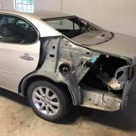 A silver sedan in a garage with its rear bumper, trunk lid, and passenger-side quarter panel removed for body repair.