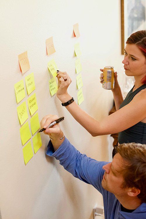 Two people are writing on sticky notes on a wall.